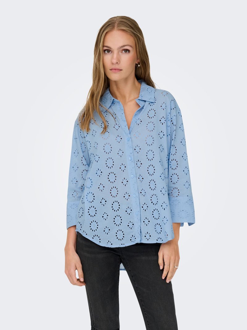 Femme portant une chemise bleu clair boutonnée avec des motifs découpés circulaires et un pantalon noir, debout devant un fond clair uni.