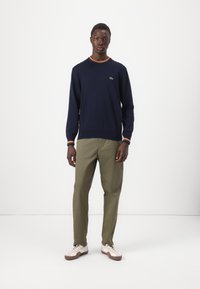 Marineblauw sweater met oranje accent bij de hals en mouwen, gecombineerd met olijfgroene broek en crèmekleurige sneakers; tentoongesteld op een effen achtergrond.