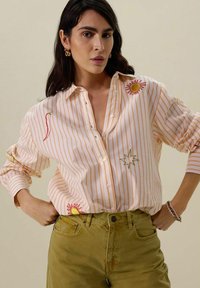 Chemise à rayures en orange pâle et blanc, ornée de broderies de soleil, d'étoile et de piments. À assortir avec un pantalon vert olive.