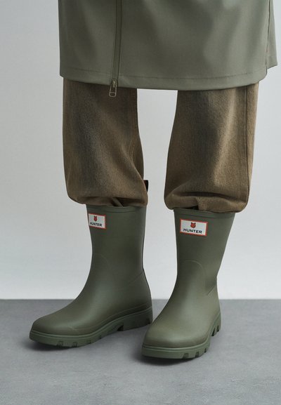 Pernas vestindo calças castanhas enfiadas em botas de chuva Hunter numa tonalidade verde-oliva, apoiadas num chão cinza, com um casaco verde-oliva correspondente parcialmente visível.