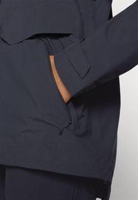 Veste navy avec une poche zippée et des poignets en velcro. Fabriquée en tissu lisse et léger, elle présente un col montant et une coupe décontractée.