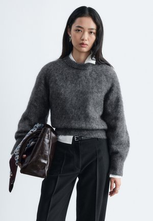 Joven mujer con el cabello largo y oscuro que lleva un suéter gris peludo y pantalones negros, sosteniendo un gran bolso de cuero marrón con una bufanda estampada.