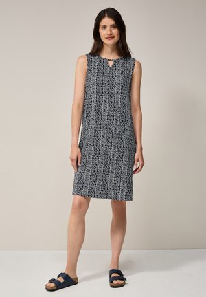 Marineblaues ärmelloses Kleid mit geometrischem weißen Muster, rundem Ausschnitt, Schlüssel-Loch-Detail an der Brust, knielang, kombiniert mit dunkelblauen Sandalen.