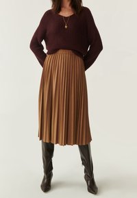 Brun plisseret midi-nederdel med en tætsiddende talje, parret med en mørkerød strikket sweater. Model bærer høje, mørkebrune hæle støvler.