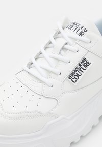 Baskets blanches avec un dessus en cuir, détails perforés, lacets plats et une semelle épaisse et texturée. Présente le logo "Versace Jeans Couture".