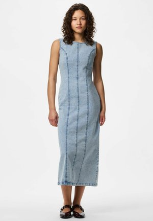 Kvinde med krøllet hår står iført en ærmeløs, lyseblå denim midi kjole og sorte Mary Jane-flade sko mod en hvid baggrund.