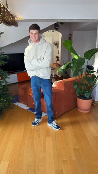 Felpa con cappuccio in pile grigio chiaro, jeans blu e sneakers blu navy. Sullo sfondo un divano arancione testurizzato e una grande pianta verde. Pavimento in legno.