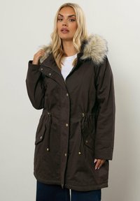 Blond kvinna i brun parkas med fuskpälskantad huva, knäppning och dragkedja fram, justerar kragen, stående mot en enfärgad bakgrund.