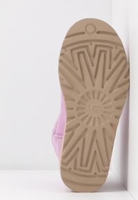 Semelle d'une botte UGG rose avec une semelle en caoutchouc beige présentant des motifs géométriques et le logo UGG au centre.