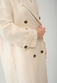 Beige uldfrakke med dobbeltknaplukning, der har store brune knapper og en spids krave. Stofret har en blød, struktureret overflade.