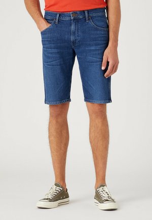 Homme portant un short en denim bleu et des baskets vert olive, debout avec une main dans la poche contre un fond uni.