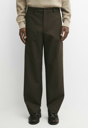Man wearing wide-leg dark brown pants, beige sweater, and black shoes, standing with hands relaxed by sides on white background.