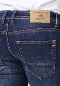 Tamno plavi traperice imaju svijetlosmeđu etiketom od kože na struku. Karakteristični crveni i bijeli naglasci na stražnjim džepovima. Detalji dvostrukog šivanja.