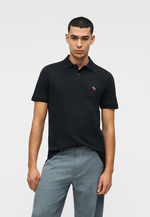 Ung mand iført en sort kortærmet poloshirt og lysegrå bukser, stående mod en ensfarvet hvid baggrund.