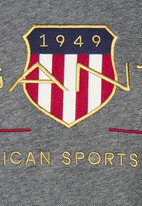 Tela gris con un escudo bordado que presenta franjas rojas y blancas, parte superior azul, borde dorado y el texto "1949" y "DEPORTES AMERICANOS."