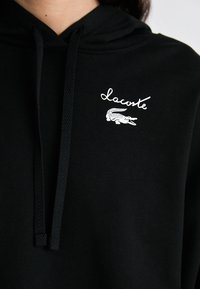 Sweat à capuche noir avec cordon de serrage, en tissu doux, et un logo brodé blanc avec un design de crocodile sur la zone gauche de la poitrine.