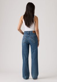 Γυναίκα με μακριά σκούρα μαλλιά, φορώντας λευκό αμάνικο τοπ και μπλε τζιν παντελόνι με φαρδιά πόδια και ψηλή μέση, γυρισμένη πλάτη σε απλό φόντο.