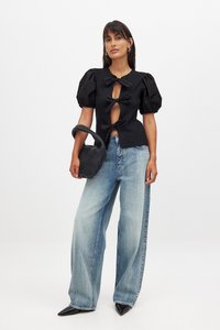 Blusa negra de mangas abullonadas con detalles de lazo y corte, combinada con jeans de pierna ancha de color azul claro y un bolso de mano negro tejido.