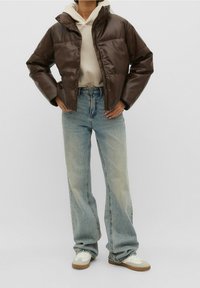 Bruine pufferjas met een rits en drukknoopsluiting, gecombineerd met een beige hoodie en lichtblauwe wijdvallende jeans. Draagt witte sneakers.