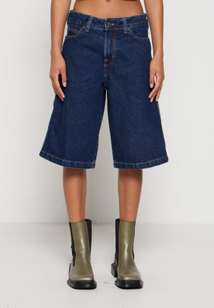 Osoba nosi tamnoplave traper hlače do koljena uparene s dvozonskim gležnjačama u maslinasto zelenoj i crnoj boji.