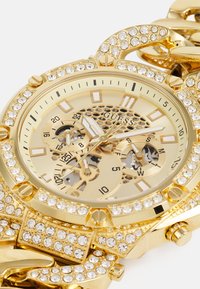 Montre en or avec un bracelet en métal brillant. Le cadran rond présente des embellissements en cristal, plusieurs cadrans et un fond à motif en nid d'abeille.