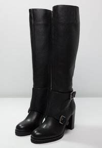 Mustat nahkaiset polven ylle ulottuvat saappaat, joissa on teksturoitu pinta, korkea muotoilu, paksut korkeat korot ja solkikoristeet nilkkaremmissä.