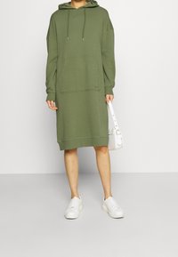 Abito a felpa color verde oliva con tasca a marsupio, polsini e orlo a coste. Abbinato a sneaker bianche e una piccola borsa bianca.