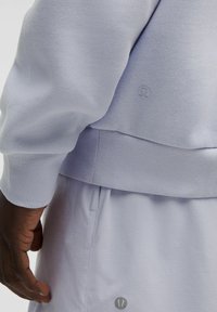 Lyse lilla sports-sweatshirt med ribbede manchetter og kant, der har et subtilt logo. Glat tekstur, lagdelt design, med et tæt billede af en hånd.
