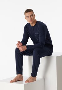 Marineblaue Langarmbluse mit Brusttasche und passender Hose, aus weichem Stoff, präsentiert an einem Modell, das auf einem Würfel sitzt.