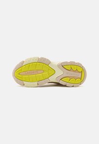A Lacoste sneaker talpa, sárga gumibetéttel, textúrált bézs külsőtalppal és kiemelkedő tapadásmintázattal. A márka logója, a 'LACOSTE' dombornyomott.