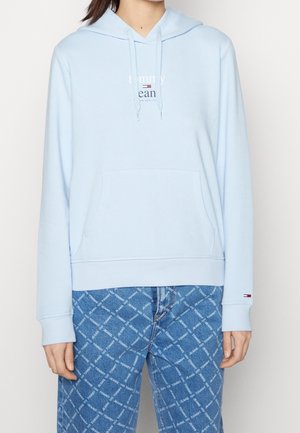 Personne portant un sweat à capuche bleu clair avec poche avant et logo, associé à un jean bleu avec un motif de texte en diagonale.