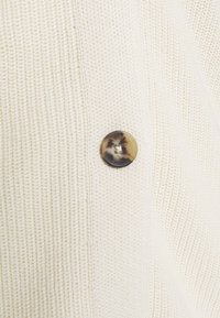 Cardigan crema lavorato a maglia con tessuto strutturato e un bottone rotondo marmorizzato con accenti scuri sul lato sinistro anteriore.