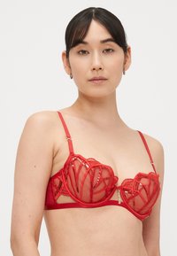 Raudonas nėriniuotas liemenėlė su skaidriomis tinklainės kaušeliais, dekoratyvine siūle ir gėlių raštu, reguliuojamomis petnešėlėmis bei auksinės spalvos metallinėmis detalėmis.