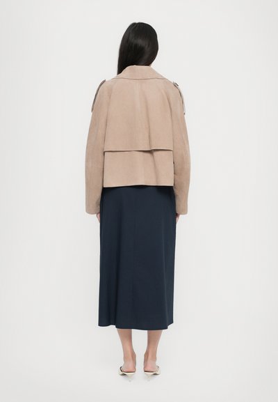 Veste en daim marron clair avec un design superposé, dotée d'un large col et de détails zippés. Portée sur une jupe bleu marine jusqu'aux chevilles.