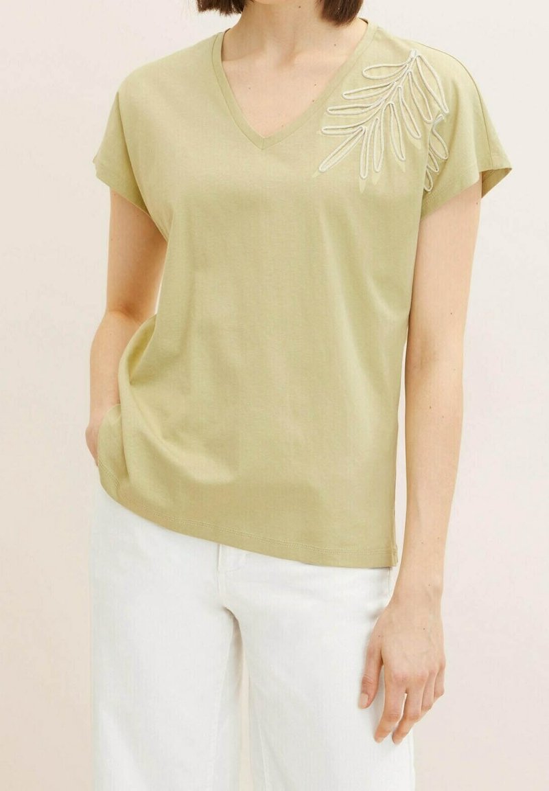 Femme portant un haut vert clair à col en V avec un motif de feuille brodé sur l'épaule et un pantalon blanc, debout avec une main dans la poche.