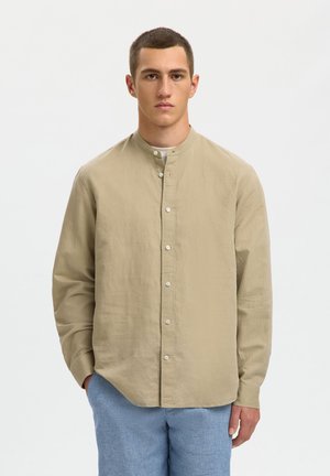 Jeune homme portant une chemise beige à col mao et un pantalon bleu, debout avec une main dans la poche contre un fond uni.