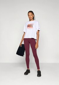 T-shirt graphique blanc à manches courtes, avec un design coloré, associée à des leggings bordeaux ajustés et des baskets noires. Une serviette sombre est tenue.