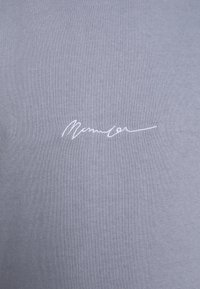 T-shirt en coton gris clair avec une signature blanche brodée discrète en bas à gauche. Texture lisse avec une coupe classique.
