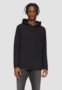 Schwarzer Kapuzenpullover mit weichem Material, langen Ärmeln und einer vorderen Kängurutasche. Das Modell trägt ihn mit grauen Jeans vor einem neutralen Hintergrund.