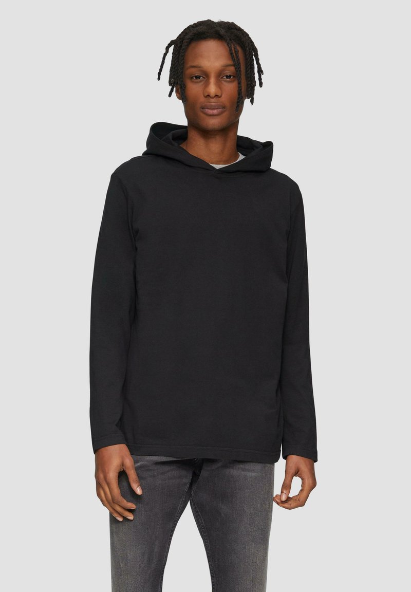 Schwarzer Kapuzenpullover mit weichem Material, langen Ärmeln und einer vorderen Kängurutasche. Das Modell trägt ihn mit grauen Jeans vor einem neutralen Hintergrund.