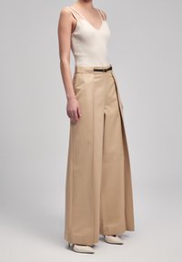 Pantalon large beige avec une texture lisse, taille haute, détail de ceinture, et un débardeur léger assorti avec des bretelles fines.