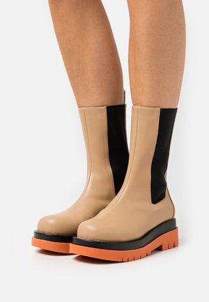 Botas bege de tornozelo com painéis laterais elásticos pretos e uma sola grossa laranja. Textura suave com um design de bico ligeiramente arredondado.