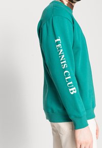 Sweatshirt turquoise à manches longues, avec "TENNIS CLUB" en blanc sur la manche gauche. Tissu doux avec des poignets et un ourlet côtelés.