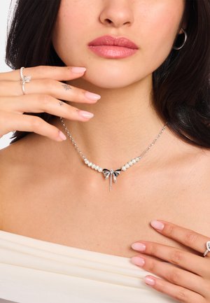 Frau trägt eine Perlenkette mit einem silbernen Schleifenanhänger, passenden silbernen Ringen und hellrosa Nagellack, berührt ihr Schlüsselbein und ihren Hals.