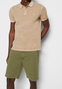 Beige pikétröja med korta ärmar, krage och subtilt logotyp. Kombinerat med olivgröna shorts som har en slät textur och rena linjer.