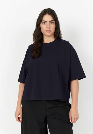 Donna con lunghi capelli scuri che indossa una camicia blu navy a maniche corte larga e pantaloni neri in piedi davanti a uno sfondo bianco uniforme.