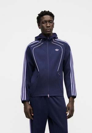 Mand iført navyblå Adidas-træningsdragt med lyse lilla striber og trefoil-logo, stående mod en ensfarvet hvid baggrund.