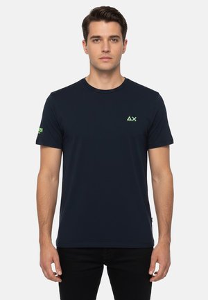 Uomo che indossa una t-shirt blu scuro tinta unita con piccoli loghi verdi sul petto sinistro e sulla manica destra, in piedi davanti a uno sfondo bianco.