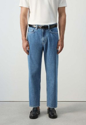 Hombre con vaqueros azul claro de pierna recta, cinturón de cuero negro, camisa blanca y mocasines negros de pie sobre suelo gris.