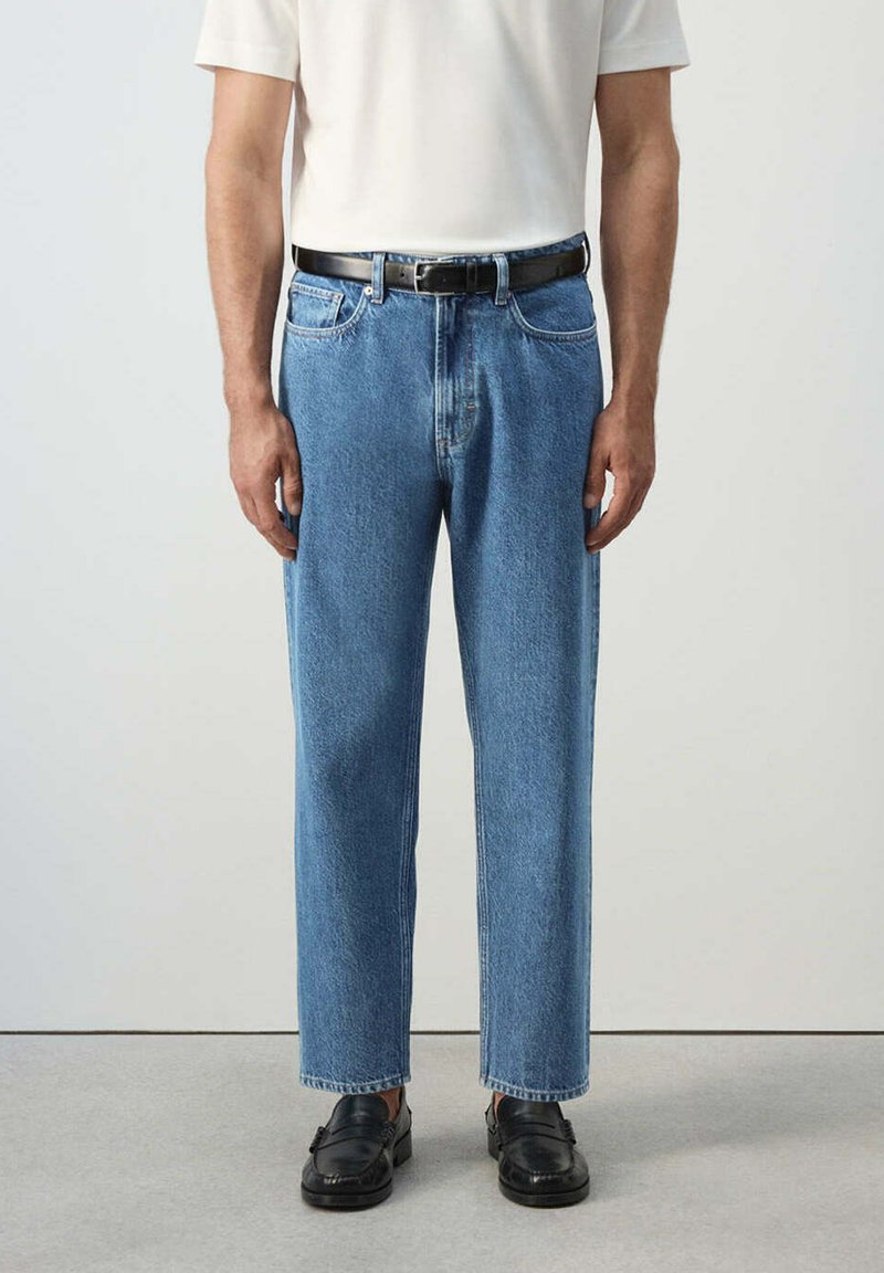 Homme portant un jean bleu clair à jambes droites, une ceinture en cuir noire, une chemise blanche et des mocassins noirs, debout sur un sol gris.